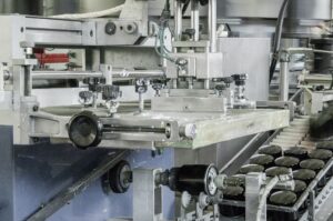 Close-up view of an industrial food processing machine, similar to those seen in MIM manufacturing, with a conveyor belt carrying jars for automated filling or packaging.